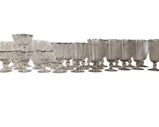231 - Silver Rim Stemware Collection Assorted Sizes (Description): A collection of clear glass stemware featuring elegant silver rim detailing. The set includes various sizes and styles of glasses, including champagne coupes, wine glasses, water goblets, and highball glasses. Each piece features a footed base and silver-toned band at the rim. Approximately 30 pieces total in varying heights and capacities.