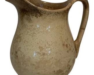 74 - Cook & Hancock Pottery Pitcher 10.5"x9.5"x6" (Description): Cook & Hancock Pottery pitcher featuring a classic form with a curved spout and loop handle. The piece displays a cream-colored glaze with characteristic brown mottling and weathered patina, giving it a rustic, antique appearance. Measures 10.5" x 9.5" x 6".