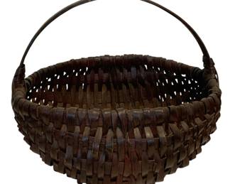 26 - Antique Woven Splint Basket w/ Handle (Description): Antique woven splint basket featuring a sturdy bentwood swing handle. The basket displays a traditional round form with a deep bowl shape, constructed of interwoven flat splints in rich brown tones showing natural patina consistent with age and use. Measures 15" x 15" x 14".