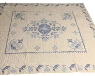 447 - Blue & White Floral Cross-Stitch Quilt 88x82" (Description): A beautiful blue and white quilt featuring intricate cross-stitch embroidery in a floral pattern. The design showcases a central medallion with a large floral rosette surrounded by leaves and smaller flower motifs. The corners feature matching floral bouquet designs, all framed by a decorative zigzag border. The outer edge is adorned with a flowing vine pattern of leaves and flowers. The quilt has an off-white or cream