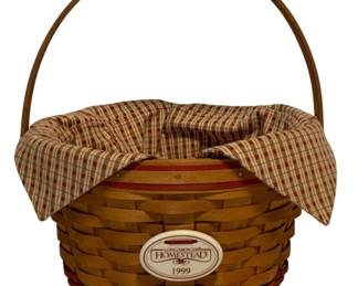 85 - Longaberger Homestead 1999 Basket w/ Liner (Description): Longaberger Homestead basket dated 1999, featuring a woven maple construction with natural and red-dipped weave accents. The basket includes its original plaid fabric liner in cream, red, and green tones. A commemorative Longaberger Homestead 1999 medallion is affixed to the front. The basket has a fixed swing handle. Measures 7 inches by 10 inches.