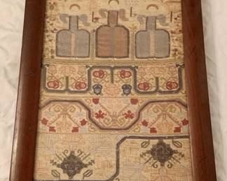 190 - Antique Framed Needlepoint Sampler (Description): Antique framed needlepoint sampler measuring 25.5 inches by 13.5 inches. This charming textile piece features traditional cross-stitch embroidery on a salmon-colored linen ground, displaying rows of alphabet letters and decorative motifs including stylized floral urns in dark blue thread. The sampler includes verses worked in multicolored threads including blue, red, green, and gold tones. Presented in a simple