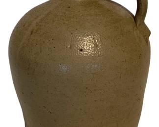 14 - Antique Salt Glazed Stoneware Jug 9"x7" (Description): Antique salt glazed stoneware jug featuring a classic ovoid form with an applied strap handle and small spout. The piece displays a warm tan/olive coloration characteristic of traditional salt glazing, with subtle speckled surface texture throughout. This utilitarian jug measures 9 inches tall by 7 inches wide.