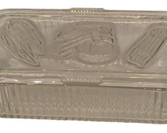 219 - Vtg Clear Glass Refrigerator Dish w/ Lid (Description): Vintage Federal Glass ribbed clear glass refrigerator dish with matching lid. Often referred to as a "fridgie". The rectangular container features ribbed sides with a decorative zigzag pattern along the base. The lid is embossed with vegetable motifs including what appears to be celery, tomatoes, and asparagus, typical of mid-century refrigerator storage dishes. Measures 8 1/2" x 4".