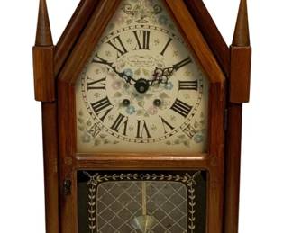 86 - New England Clock 8-Day Mantle Clock w/ Key (Description): New England Clock Company 8-day mantle clock with key. This steeple-style clock features a Gothic-inspired wooden case with pointed finials flanking the peaked top. The clock face displays Roman numerals on a decorative floral dial with ornate hands. The lower glass panel reveals a brass pendulum and features gold-toned lattice pattern with leaf motif border. The case has a warm medium wood finish. Measures 11" x 22" x 6".