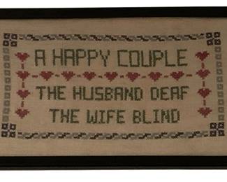 6 - Framed Needlepoint "A Happy Couple" Sampler (Description): Framed needlepoint sampler featuring a humorous sentiment reading "A Happy Couple / The Husband Deaf / The Wife Blind." The cross-stitch piece is worked in muted colors including burgundy, pink, and sage green, with decorative heart motifs and geometric borders on cream fabric. Presented in a black wooden frame. Measures 8.5" x 13.75".