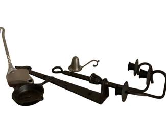 227 - Wrought Iron Candle Holders & Snuffer (Description): A collection of wrought iron and metal candle accessories including wall-mounted candle holders with curved arms and drip pans, a candle snuffer with bell-shaped head, a small handled dish or chamberstick, and a decorative strap hinge or bar. The pieces feature traditional blacksmith-style craftsmanship with twisted details and hand-forged elements, suited for rustic or colonial decor.