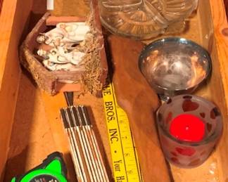 56 - Drawer Lot of Assorted Items (Description): A miscellaneous drawer lot containing a small nativity scene with ceramic figures in a cork or bark manger, a clear pressed glass dish with scalloped edges, a silver-toned metal bowl, a decorative glass votive holder with red hearts containing a red candle, a set of metal fondue forks or skewers, a tape measure, and a carpenter's folding ruler. An assorted collection of household items.
