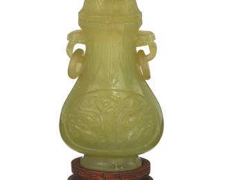 1304 A Chinese carved jade lidded vessel 20th century Carved with Taotie mask to body, flanked by handles with ring accents and a floriform finial, raised on a hardwood stand Jade: 6" H x 3" W x 1.875" D; With stand: 7.25" H Estimate: $300 - $500