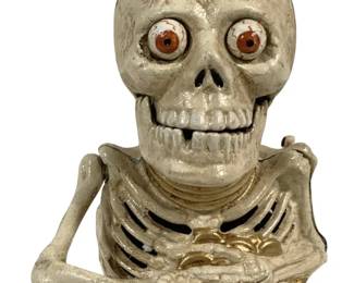 1520 - Cast Iron Skeleton Bank 7"x5.5"x6" (Description): Cast Iron Skeleton Bank featuring a  half-figure skeleton with expressive glass  eyes and articulated arms. The piece is  painted in an antiqued cream finish with gold  accents visible on the interior ribcage and hands. This mechanical bank depicts a  skeleton bust with detailed bone structure  including skull, ribcage, and arms positioned  to hold coins. Measures 7" x 5.5" x 6".