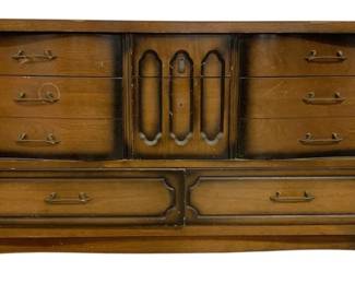 7593 - Mid-Century 8 Drawer Dresser 31"x60"x18" (Description): Mid-century style eight drawer dresser  featuring warm walnut-toned wood finish. The  dresser showcases a center cabinet with  decorative carved door panels flanked by six  side drawers with brass-tone pulls. Two large bottom drawers with matching hardware and  carved frame details provide additional  storage. Shows vintage wear consistent with  age. Measures 31 inches high by 60 inches  wide by 18 inches deep.
