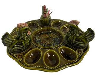 1549 - Mid-Century Green Ceramic Deviled Egg Set (Description): Mid-Century deviled egg serving set in olive  green glazed ceramic, consisting of four  pieces. The set includes a large round egg  plate with twelve indentations for deviled  eggs, featuring a decorative center medallion with a rooster design surrounded by floral  accents and dotted border details.  Accompanying the plate are three figural  chicken pieces, including one larger rooster  and two smaller hen-shaped pieces, likely