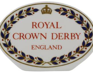 799 - Royal Crown Derby England Display Plaque 3" (Description): Royal Crown Derby display plaque featuring  the company's signature branding. The oval  porcelain plaque displays "Royal Crown Derby  England" in red lettering, topped with a gold  crown. A decorative border of cobalt blue leaves with red berries and gold accents  encircles the text. Measures 3 inches.