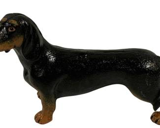 1514 - Cast Iron Dachshund Dog Figure 10"x7"x3" (Description): A charming cast iron dachshund dog figure,  hand-painted in classic black and tan  coloring. The figurine captures the  distinctive long body and short legs  characteristic of the breed, with detailed facial features including expressive eyes and  tan markings on the muzzle, eyebrows, and  paws. The piece has a glossy finish and would  serve well as a doorstop or decorative  accent. Measures 10" x 7" x 3".