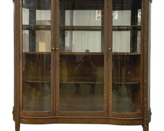 7504 - Antique Oak Curved Glass China Cabinet (Description): Antique oak china cabinet featuring elegant  curved glass panels on the front and sides.  This handsome display piece showcases  beautiful quarter-sawn oak with distinctive  tiger oak grain pattern throughout. The cabinet features double doors with brass  hardware, multiple interior shelves for  display, and rests on tapered legs with  caster wheels for easy movement. The  serpentine front design and quality