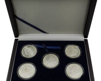 810 - Boxed Set of 5 Morgan Silver Dollars 1881-1889 (Description): A boxset containing five Morgan Silver  Dollars presented in individual protective  capsules within a velvet-lined presentation  case. The collection includes coins dated  1881-S, 1884-O, 1885-O, 1887, and 1880-S, representing various mint marks from San  Francisco and New Orleans. The dark wooden  presentation box features a hinged lid with  ribbon closure and navy blue velvet interior  display tray.