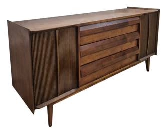 1413 - Lane First Edition Mid-Century Credenza (Description): Lane credenza in walnut featuring mid-century  modern design. The piece showcases three  center drawers with louvered fronts flanked  by two side cabinets with vertical ribbed  tambour doors. The credenza rests on tapered peg legs characteristic of the period.  Measures 31 inches high by 66 inches wide by  18 inches deep.