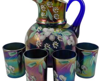 737 - Fenton Cobalt Blue Cherry Blossom Pitcher (Description): Vintage Fenton cobalt blue carnival glass  pitcher set featuring a hand-painted cherry  blossom design. The set includes a large  pitcher with a cobalt blue applied handle and  four matching tumblers. The iridescent carnival glass finish displays beautiful  rainbow hues of purple, gold, and green. Each  piece is decorated with white cherry  blossoms and green foliage. Pitcher measures  10 inches tall.