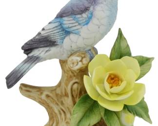 1072 - Andrea by Sadek Mountain Bluebird Figure (Description): Andrea by Sadek porcelain figure depicting a  Mountain Bluebird perched on a naturalistic  tree stump base. The bird features delicate  hand-painted plumage in shades of blue,  white, and purple-gray. The base is adorned with a yellow rose bloom and green leaves.  Measures 6 inches tall.