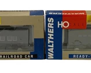 1584 - Walthers HO Gold Line Military Train Cars (Description): Two Walthers Gold Line HO scale military  train cars. These ready-to-run model railroad  cars feature gray military-style paint  schemes and come in their original Walthers  packaging. The set includes two boxed cars from the Gold Line series, designed for HO  scale layouts.