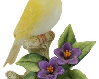 1082 - Andrea by Sadek Porcelain Canary Figure 5.5" (Description): Andrea by Sadek porcelain canary figure  depicting a yellow canary perched on a  naturalistic tree stump base adorned with  purple flowers and green foliage. The bird is  finely detailed with realistic plumage in shades of yellow and cream. Measures  5.5inches tall.