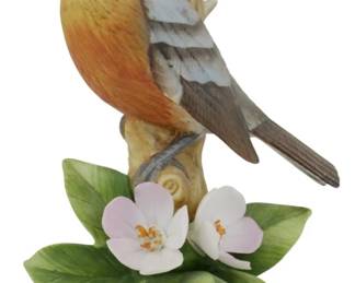 1086 - Andrea by Sadek Porcelain Robin Figure 6.5" (Description): Andrea by Sadek porcelain robin figure. This  charming hand-painted figurine depicts an  American robin perched on a naturalistic tree  stump base adorned with delicate pink apple  blossoms and green leaves. The bird features realistic coloring with anorange-red breast,  brown and gray wing feathers, and yellow  beak. Measures 6.5 inches tall.