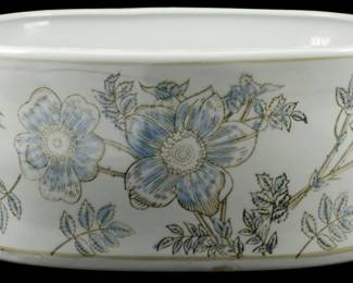 1027 - Blue Flower Decorated Planter 6"x13"x9" (Description): White ceramic planter featuring a decorative  blue floral motif with gold accents. The  oval-shaped vessel displays large stylized  flowers and delicate foliage wrapping around  the exterior. The planter is fitted with small handles on each end and sits on a  footed base. Measures 6 inches high by 13  inches long by 9 inches wide.