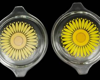 1231 - 2 Pyrex Sunflower Lids (Description): Two Pyrex sunflower lids featuring clear  glass with yellow sunflower designs on the  underside. Each lid has tab handles on  opposite sides for easy gripping. The  sunflower patterns show radiating yellow petals surrounding circular centers, with  smalldecorative flower accents around the  perimeter.