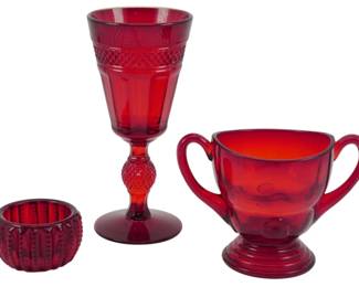 761 - 3 Assorted Ruby Glass Items (Description): A collection of three assorted ruby glass  items including a tall footed goblet with  diamond point pattern detailing on the stem  and decorative banding, a two-handled sugar  bowl on a pedestal base, and a small ribbed votive holder or salt cellar. All pieces  feature deep red coloring characteristic of  ruby glass.