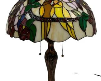 1510 - Tiffany Style Stained Glass Table Lamp 24" (Description): Tiffany style stained glass table lamp  featuring a colorful dome shade with a pair  of yellow birds perched on a branch amid pink  flowers, green foliage, and purple grapes.  The scalloped edge shade displays intricate leaded glass work in cream, gold,green, pink,  blue, and burgundy tones. The lamp sits on a  cast metal base with a bronze-tone finish in  an Art Nouveau style with decorative leaf  motifs. Features dual pull chain operation.