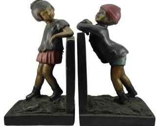 1255 - Pair Bronze Boy & Girl Large Bookends 11" (Description): Pair of bronze bookends featuring charming  figural sculptures of a boy and girl. The  girl is depicted wearing a gray top with a  red pleated skirt, her hair pulled back in a  bun, while the boy wears a red cap, dark long-sleeved shirt, and green shorts. Both  figures lean playfully against the vertical  book supports in animated poses. The  sculptures feature polychrome patina  detailing and rest on textured rectangular