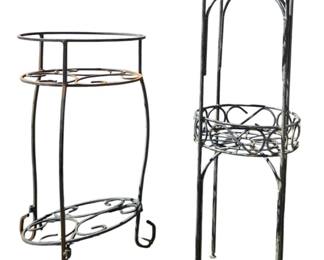 7697 - 2 Metal Plant Stands 28"x10.5" (Description): Two metal plant stands featuring decorative  scrollwork designs. The stands are finished  in black with a distressed patina, each  featuring curved legs and ornamental wire  detailing. One stand is taller with a two-tier design, while the other is shorter  with a single display tier. Measures 28  inches by 10.5 inches.