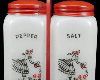 1137 - Pr McKee Watering Can Lady Shakers 4.5" (Description): Pair of McKee Watering Can Lady salt and  pepper shakers measuring 4.5 inches tall. The  shakers feature white milk glass bodies with  red painted metal lids and sit in a matching  red metal carrying caddy with center handle. Each shaker is decorated with a charming  transfer design depicting a lady in a full  skirt watering flowers, rendered in black  with red accents. The word "PEPPER" appears  on one shaker and "SALT" on the other.