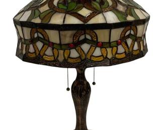 1507 - Tiffany Style Stained Glass Table Lamp 24" (Description): Tiffany style stained glass table lamp  featuring a domed shade with an intricate Art  Nouveau-inspired design. The shade displays  a colorful pattern of interlocking rings and  flowing motifs in shades of amber, green, cream, and burgundy, accented with red  jeweled cabochons. The shade rests on a cast  metal base with a bronze-tone finish,  featuring a ribbed stem and decorative floral  base. The lamp operates with dual pull