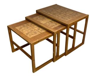 1555 - Tile Top Mid-Century Wood Nesting Tables (Description): Set of three wooden nesting tables featuring  decorative tile inlay tops with floral  botanical motifs. The tables are crafted in a  warm medium-tone wood with clean mid-century  modern lines and angular stretcher bases. Each table features matching ceramic tiles  with impressed leaf and flower designs in  earth tones. The largest table measures 23.5  inches wide by 16 inches deep by 18.5 inches  tall, with the two smaller tables designed to