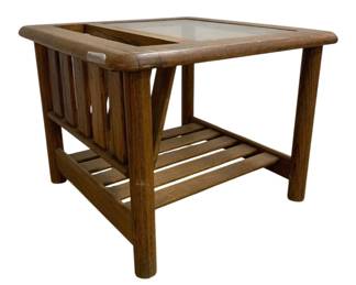 7614 - Mid-Century Oak Mersman Table w/ Smokey Glass (Description): Mid-century style oak end table featuring a  built-in magazine rack on one side. The table  showcases craftsman details including a  glass inset top, slatted lower shelf, and  sturdy round legs. The warm oak finish highlights the natural wood grain throughout.  Measures 20 inches tall by 26 inches wide by  26 inches deep.