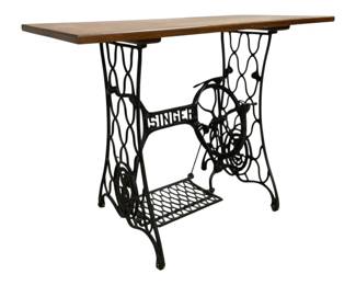 7675 - Singer Sewing Table 36"x40"x21" (Description): Singer sewing table featuring an antique cast  iron treadle base with the iconic Singer  name prominently displayed. The ornate black  iron base includes the original foot pedal  and decorative scrollwork design typical of vintage Singer machines. Topped with a  warm-toned wooden surface, this piece has  been repurposed as a functional table.  Measures 36"x40"x21".