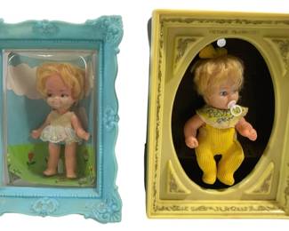 2014 - 2 Vintage Picture Playmates Dolls in Frames (Description): Two vintage dolls displayed in decorative  plastic picture frames. One frame is  turquoise blue with ornate baroque-style  molding, featuring a blonde doll in a white  dress with a painted floral background. The second frame is yellow with scroll detailing,  marked "Picture Playmates," containing a  blonde baby doll wearing yellow corduroy  overalls with a decorative bib. Each frame  measures 8" x 6".