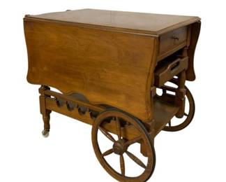 7503 - Vintage Wooden Drop Leaf Tea Cart w/ Wheels (Description): Vintage wooden tea cart featuring a drop leaf  design with turned legs and decorative spoke  wheels. The cart includes a single drawer  with brass knob pull and a removable serving  tray beneath. A lower shelf provides additional storage space. The piece displays  warm honey-toned wood finish in traditional  Early American style. Measures 28" x 46" x  34".
