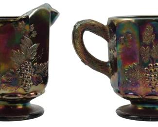 718 - Cobalt Blue Grape Leaf Carnival Glass Set (Description): Vintage cobalt blue carnival glass creamer  and sugar set featuring an embossed grape  leaf and grape cluster design. The pieces  display the characteristic iridescent rainbow  finish typical of carnival glass, with rich purple, green, and gold tones shimmering  across the surface. Each piece features  textured rope-style handles and sits on a  footed base. Measures 4 inches.