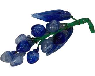 2010 - Blue & Clear Art Glass Grape Cluster Decor 7" (Description): Decorative blue and clear art glass grape  cluster featuring an assortment of glass  grapes in varying shades of cobalt and light  blue, accented with blue glass leaves, all  attached to a green painted metal stem. The piece displays Murano-style craftsmanship  with textured surfaces on the individual  grape forms. Measures 7 inches long.