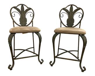 7629 - Pair of Metal Pineapple Back Bar Stools (Description): Pair counter height bar chairs featuring  decorative pineapple and palm leaf motifs on  the heart-shaped backs. The stools have  curved scrollwork legs with an X-stretcher  base and acanthus leaf detailing. Each chair has an upholstered cushion seat in a neutral  beige tone. The metal has a verdigris patina  finish. Measurements: 41"x17"x17" with a 24"  seat height.