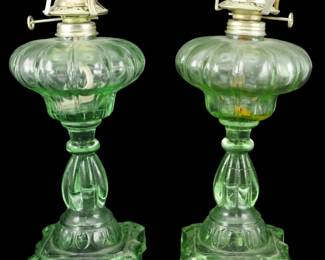 1245 - Vtg Pair Green Pressed Glass Oil Lamps (Description): Vintage pair of green pressed glass oil lamps  featuring melon-ribbed fonts atop decorative  pedestal bases with scalloped edges. Each  lamp retains its original burner hardware  with wick adjusters. The uranium-style green glass displays an elegant translucent quality  with a classic Victorian aesthetic. Measures  11.5 inches tall.