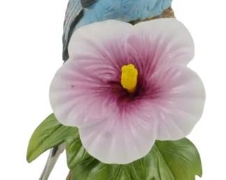 1085 - Andrea by Sadek Porcelain Parakeet Figure (Description): Andrea by Sadek porcelain figurine depicting  a blue parakeet perched on a naturalistic  branch with a pink and white hibiscus flower  and green leaves. The bird features detailed  hand-painted plumage in shades of blue, white, and gray with realistic feather  texturing. Measures 6.5 inches tall.