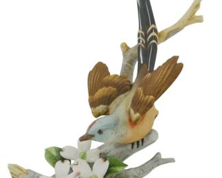 1078 - Andrea by Sadek Scissor-Tailed Flycatcher (Description): Andrea by Sadek porcelain Scissor-Tailed  Flycatcher figure. This beautifully crafted  piece depicts the graceful bird perched on a  naturalistic branch base with white dogwood  blossoms and green foliage. The flycatcher is rendered with exceptional detail, featuring  its distinctive long tail feathers in gray  and white, warm brown wings, pale blue-gray  head, and subtle peachy-orange accents on the  breast. The figure measures 10 inches tall.
