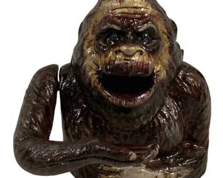 1513 - Cast Iron Bigfoot Bank 7"x6"x6" (Description): Cast iron Bigfoot bank featuring a detailed  sculpture of the mythical creature in a  seated pose with open mouth serving as the  coin slot. The piece is hand-painted in rich  brown tones with tan highlights accentuating the facial features and fur texture. The  figure has expressive blue eyes and  outstretched arms forming a tray-like base.  Measures 7"x6"x6".