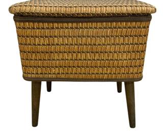7590 - Mid-Century Woven Sewing Basket Stand (Description): Mid-century style woven rattan storage basket  featuring a hinged lid and tapered wooden  legs. The basket displays a natural woven  pattern with contrasting darker accents along  the edges. The design combines functional storage with period-appropriate aesthetics.  Measures 18 inches by 16 inches by 11 inches.