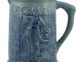 743 - Vtg Stoneware Blue & White Bar Scene Pitcher (Description): Vintage stoneware pitcher featuring a blue  and white glaze with a molded bar scene in  relief. The design depicts a tavern keeper or  barmaid figure in period dress, holding up a  drink, set against an architectural backdrop with decorative elements. The handle  features a textured rope-like pattern.  Measures 9 inches tall.