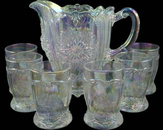 740 - Mosser Clear Iridescent Dahlia Pitcher Set (Description): Vintage Mosser clear iridescent Dahlia  pattern glass pitcher set. The set features a  large pitcher with ornate scalloped rim and  decorative handle, accompanied by six  matching footed tumblers. Each piece displays the distinctive Dahlia floral motifin  pressed glass with a beautiful rainbow  iridescent finish. Pitcher measures 8 inches  tall.