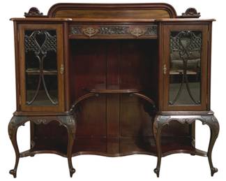 7760 - English Victorian Mahogany Display Cabinet (Description): Victorian mahogany display cabinet featuring  an elegant serpentine form with carved  decorative elements. The piece includes two  flanking glass-fronted side cabinets with  ornate fretwork overlays and interior shelving, flanking a central open display  area with a curved shelf. The cabinet is  adorned with carved foliate details along the  frieze and apron, decorative brass hardware  and escutcheons, and stands on graceful
